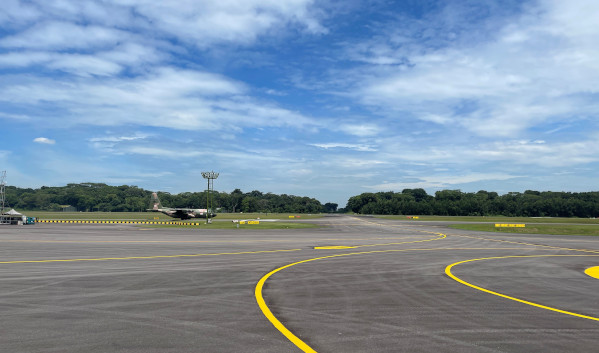Airport runway view with blue sky, green trees, and a military plane parked on the side.